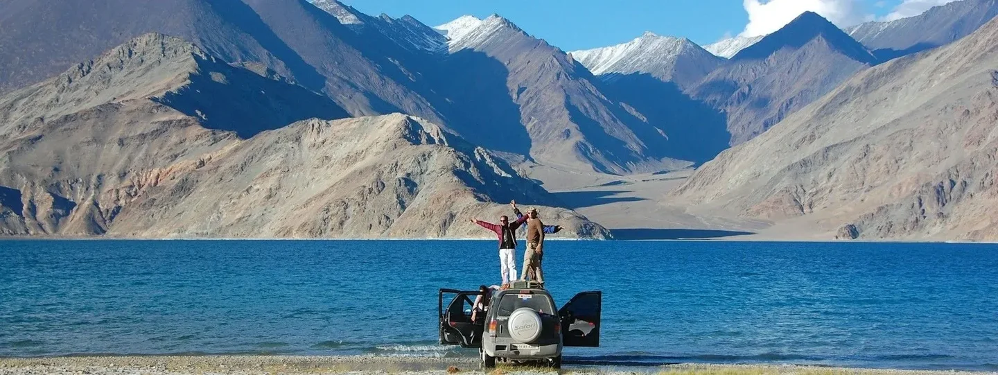 People standing beside an SUV overlooking mountain and ocean landscape during a scenic road trip.